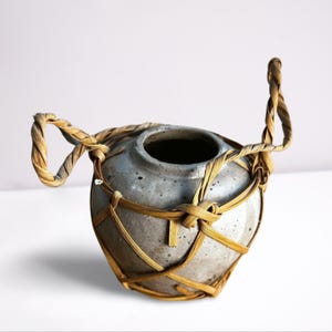 Pode incluir: Um pequeno vaso de cerâmica cinza com uma alça de palha trançada e uma cesta de palha trançada ao redor do corpo.