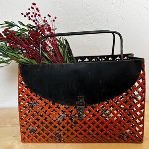 Puede incluir: Una cesta decorativa de metal con asa y solapa superior negras. El cuerpo de la cesta es un diseño de celosía en naranja, con un cierre de metal. La cesta está llena de bayas rojas y follaje verde.
