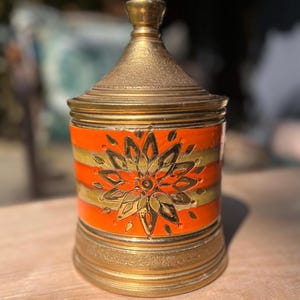 Op de afbeelding: Een decoratieve pot met een gouden deksel en basis. De body van de pot is oranje met gouden horizontale strepen en een gouden bloemenontwerp. De pot heeft een conisch deksel en een cilindrisch lichaam.