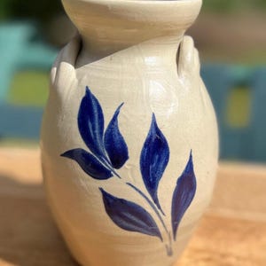 May include: A cream-colored ceramic vase with a flared rim and two small side handles. The body of the vase features a hand-painted cobalt blue leaf motif consisting of five distinct leaves arranged on a single stem, centered on the front surface.