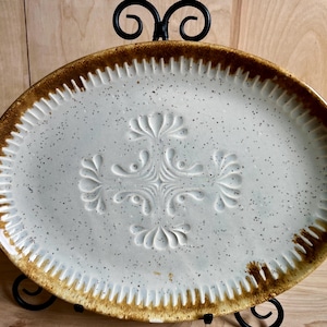 May include: Oval ceramic serving platter with a speckled white surface and a raised floral design in the center. The rim is a warm brown with a textured, ridged edge. The platter is displayed on a black metal stand.