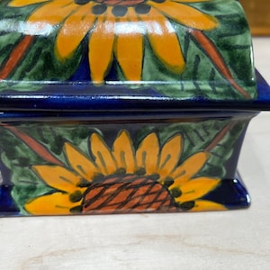 Puede incluir: Una caja rectangular de cerámica con tapa, que presenta un diseño de girasol pintado a mano. La caja tiene un acabado brillante con pétalos amarillos vibrantes, un centro marrón y hojas verdes sobre un fondo azul profundo en la base.