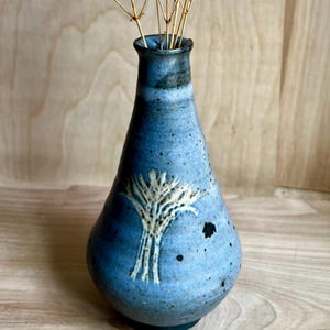 May include: A blue ceramic vase with a tree design. The vase has a narrow neck and a wider body, with a white tree design on the front. The vase is filled with dried stems. The vase is approximately 8 inches tall.