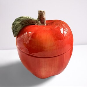 Puede incluir: Un recipiente de cerámica rojo en forma de manzana con una hoja verde en la parte superior. El recipiente tiene un acabado brillante.