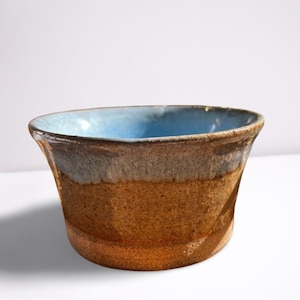 May include: A handmade ceramic bowl with a light blue glaze inside and a brown glaze on the outside. The bowl has a slightly flared rim.