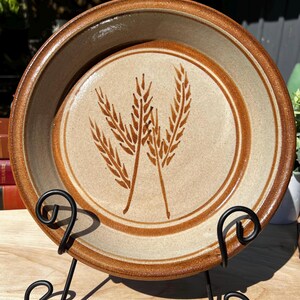 Puede incluir: Un juego de dos platos de cerámica con un diseño de espigas de trigo. Los platos son de color beige con bordes marrones e ilustraciones de trigo. Se exhiben en un soporte de metal negro, adecuado para la decoración de la cocina o el comedor.