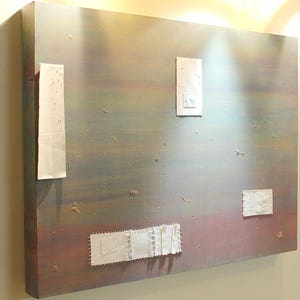 Puede incluir: Obra de arte abstracta sobre un marco de madera. La pieza presenta una mezcla de colores, incluyendo marrón, gris y toques de azul y rojo. Varios parches rectangulares de tela blanca están adheridos a la superficie.