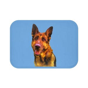 May include: A bath mat featuring a German Shepherd dog with brown and black fur, set against a light blue background. The dog's head is centered, with its mouth slightly open. The mat has rounded corners.