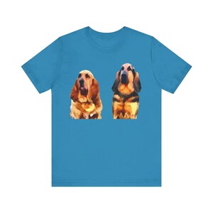 May include: A turquoise t-shirt featuring a watercolor-style illustration of two Basset Hound dogs. The dogs are depicted in shades of brown, tan, and black, with long ears and droopy faces. The shirt is made of a soft material.