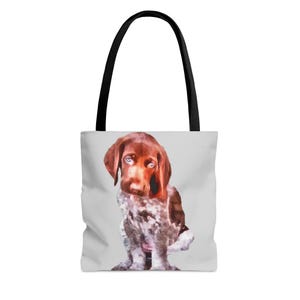 Puede incluir: Un bolso tote con una ilustración de cachorro al estilo acuarela. El cachorro tiene pelaje marrón con manchas blancas y marrones y ojos azules. El bolso tiene un asa negra y un fondo gris claro.