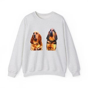 Puede incluir: Una sudadera gris claro con dos ilustraciones de perros Basset Hound al estilo acuarela. Los perros están representados en tonos marrones, negros y beige. La sudadera tiene cuello redondo y mangas largas.