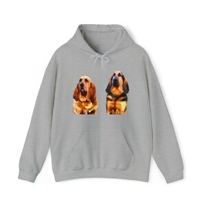Puede incluir: Una sudadera con capucha gris claro con una ilustración al estilo acuarela de dos perros Basset Hound. Los perros están representados en tonos marrones, negros y beige. La sudadera tiene un bolsillo delantero y una capucha con cordón.