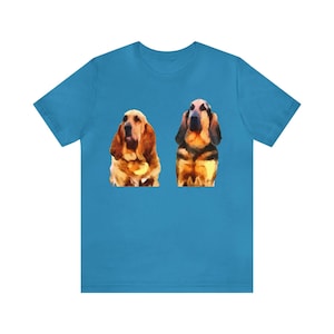 May include: A turquoise t-shirt featuring a watercolor-style illustration of two Basset Hound dogs. The dogs are depicted in shades of brown, tan, and black, with long ears and droopy faces. The shirt is made of a soft material.
