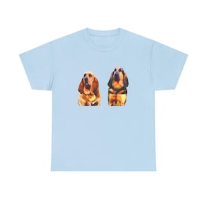 Puede incluir: Camiseta azul claro con dos ilustraciones de perros Bloodhound al estilo acuarela. Los perros están representados en tonos marrones, negros y beige. La camiseta tiene cuello redondo clásico y mangas cortas.