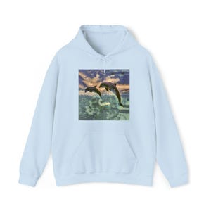 Könnte beinhalten: Hellblauer Kapuzenpullover mit einer Grafik von zwei Delfinen, die aus dem Meer springen, vor einem Sonnenuntergang. Die Delfine sind dunkelgrau, und das Wasser ist eine Mischung aus Blau und Grün. Der Hoodie hat eine Vordertasche und eine Kapuze.