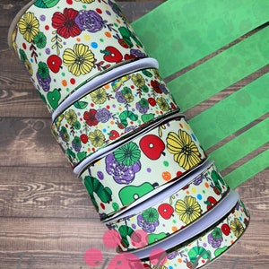 May include: A stack of five rolls of green and white ribbon with a colorful pattern of flowers, apples, and caterpillars. The ribbon is 2.5 inches wide.