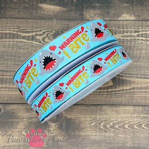 May include: Three rolls of blue ribbon with a white shark graphic and the text "Warning I Bite" in gold glitter. The shark has red teeth and hearts around it.