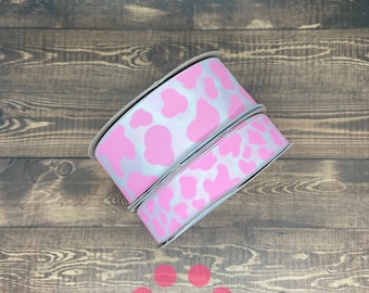 May include: Two rolls of white and pink ribbon with a paw print pattern. The ribbon is on a wooden surface with the text "Pawsitively Cute" printed below.