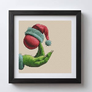 May include: A framed cross-stitch art piece featuring a green hand holding a red ornament topped with a Santa hat. The artwork has a beige background and is framed in a black frame. The image is a holiday-themed design.