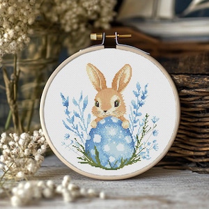 Könnte beinhalten: Ein Kreuzstich-Stickrahmen mit einem blauen und weißen Osterei und einem braunen Hasen, der das Ei hält. Der Hase ist von blauen und weißen Blumen umgeben.