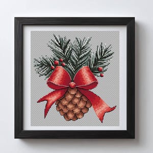 May include: Cross-stitch artwork of a pinecone with a red bow and evergreen branches. The pinecone is brown, the bow is red, and the branches are green with red berries. The artwork is framed in a black frame.
