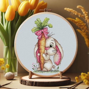 May include: A cross-stitch embroidery hoop featuring a cartoon bunny hugging a large carrot with a pink bow. The bunny has pink-lined ears and a green-eyed expression. The background is light blue, and the hoop is displayed on a wooden stand.