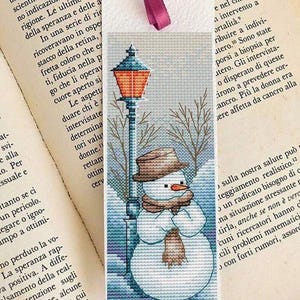 Puede incluir: Marcapáginas de punto de cruz con un muñeco de nieve en una escena invernal con una farola. El muñeco de nieve lleva un sombrero y una bufanda, con un fondo nevado. Se adjunta una cinta burdeos. El marcapáginas está sobre un libro.