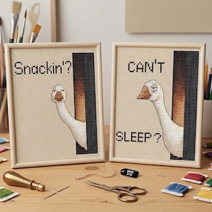 May include: Two framed cross-stitch artworks featuring white geese peeking from behind a black vertical panel. The left piece displays the text "Snackin'?" and the right piece displays the text "CAN'T SLEEP?" in black stitched lettering.