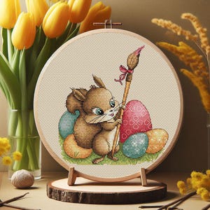 May include: A cross-stitch embroidery hoop featuring a brown squirrel holding a paintbrush with a pink bow. The squirrel is surrounded by colorful Easter eggs. The embroidery is displayed on a wooden stand, with yellow tulips in the background.