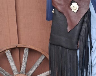 May include: A handmade leather crossbody bag with a brown flap and black body, adorned with long black fringe. The flap features a decorative bone button. The bag is suspended by a black cord.