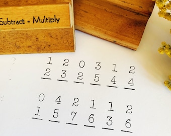 May include: A close-up of a white sheet of paper with handwritten math problems. The problems are written in black ink and include the numbers 1, 2, 0, 3, 5, 4, 7, 6, and 1. The paper is surrounded by two wooden stamps. One stamp says "Subtract - Multiply" and the other says "Divide No. 5".
