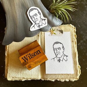 Könnte beinhalten: Ein Holz-Stempel mit dem Wort "Wilson" darauf gedruckt. Der Stempel liegt auf einem Stück weißem Stoff mit einem dekorativen Rand. Auf dem Stoff befinden sich zwei Schwarzweißzeichnungen von Männern.