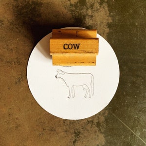 May include: A wooden stamp with the word "COW" printed on it. The stamp is on a white circular piece of paper with a line drawing of a cow.