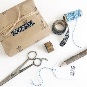 May include: A brown paper parcel wrapped with twine and decorated with a floral patterned tape. The parcel is sitting on a white surface with a wooden stamp that says "whe", a roll of blue and white twine, a roll of floral patterned tape, a pair of scissors, a twig with a pencil attached, and a white tag with a stamped image of wheat.