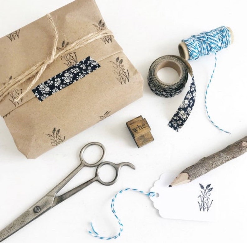May include: A brown paper parcel wrapped with twine and decorated with a floral patterned tape. The parcel is sitting on a white surface with a wooden stamp that says "whe", a roll of blue and white twine, a roll of floral patterned tape, a pair of scissors, a twig with a pencil attached, and a white tag with a stamped image of wheat.