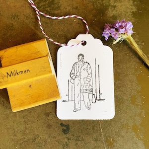 May include: A white tag with a black and white illustration of a person wearing a coat and holding a bag. The tag is tied with a red and white string. The text "Milkman" is printed on a wooden block.