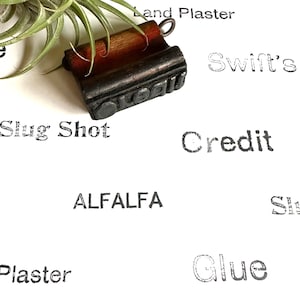 May include: A close-up of a vintage wooden stamp with the word "Oval" embossed on it. The stamp is sitting on a white surface with other words printed around it, including "Slug Shot", "Credit", "Land Plaster", "Swift's", "ALFALFA", "Glue", and "Plaster".