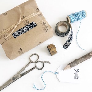 May include: A selection of craft items on a white background. Features a wrapped parcel with twine, floral tape, blue and white string, scissors, a wooden stamp, a pencil, and a blank tag with a floral design.