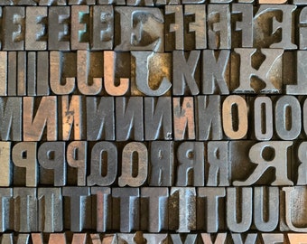 May include: A close-up of a collection of vintage letterpress printing blocks, arranged in rows. The blocks are made of wood and metal, and they feature a variety of letters, numbers, and symbols. The blocks are all slightly worn and have a rustic, antique look.