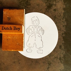 May include: A wooden rubber stamp with the text "Dutch Boy" on it. The stamp is used to create a design of a boy wearing a hat and overalls.