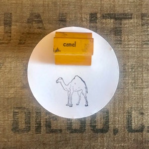 Puede incluir: Un sello de goma amarillo con la palabra "camel" impresa en él. El sello está sobre una superficie circular blanca con un dibujo de línea negra de un camello.