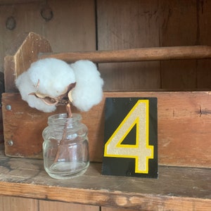 May include: A small, clear glass jar with a cotton stem and a black sign with a gold glitter number 4. The items are displayed on a wooden shelf with a wooden toolbox in the background.