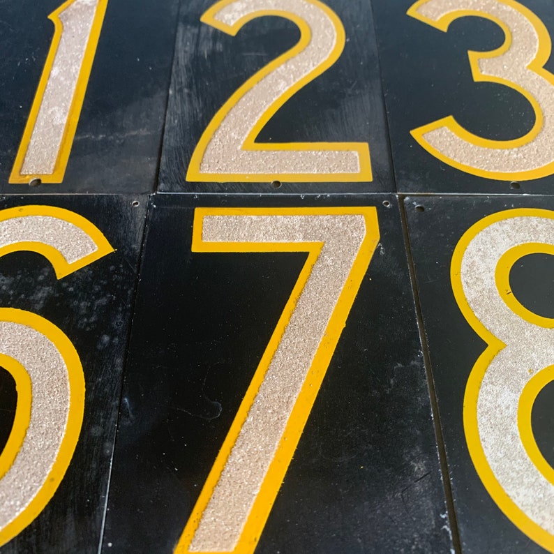 May include: Close-up of several black metal number tiles. The numbers 1, 2, 3, 6, 7, and 8 are visible. Each number has a silver glitter-like texture with a yellow border. The numbers are on a black background.