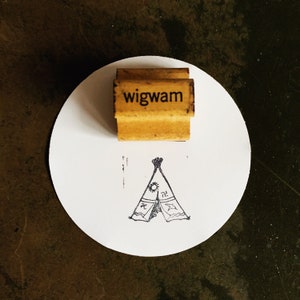 May include: A wooden stamp with the word "wigwam" in black lettering. Beneath the stamp is a drawing of a teepee with a sun, swastika, and other symbols. The stamp and drawing are on a white circle.