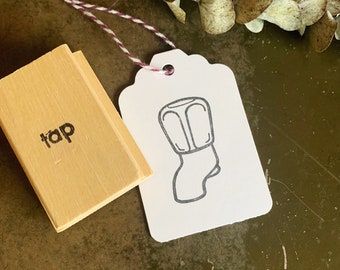 May include: A wooden stamp with the word "tap" printed on it. The stamp is next to a white tag with a black and white illustration of a tap.