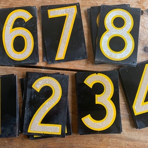 May include: A set of black metal number signs, each displaying a silver glitter number with a yellow border. The numbers 1 to 8 are present, with a small hole at the top for hanging. These signs could be used for house numbers or other forms of identification.