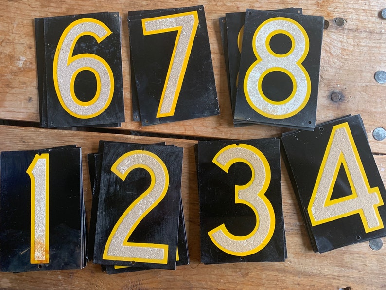 May include: A set of black metal number signs, each displaying a silver glitter number with a yellow border. The numbers 1 to 8 are present, with a small hole at the top for hanging. These signs could be used for house numbers or other forms of identification.