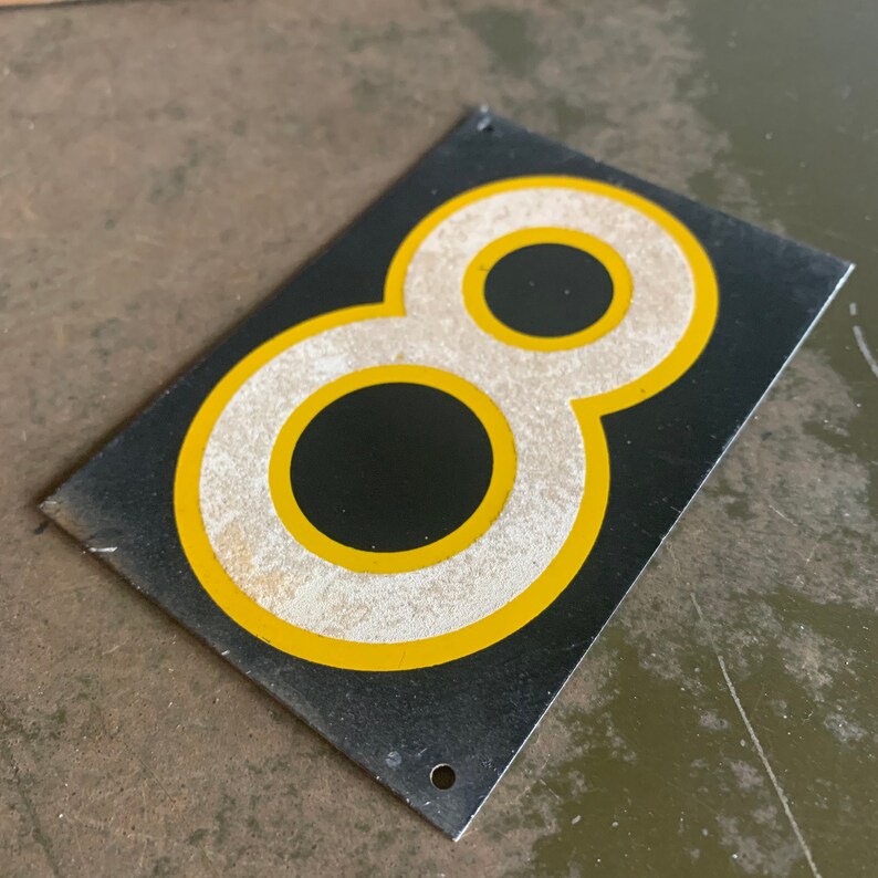 May include: A rectangular black metal sign displaying the number eight. The number is white with a yellow border. The sign has two mounting holes and shows signs of wear and tear.