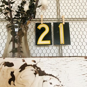 May include: Two black rectangular signs displaying the numbers 2 and 1 in yellow, suspended against a wire mesh backdrop. The numbers have a distressed, vintage aesthetic. A clear glass bottle with dried branches rests on a white, distressed shelf.
