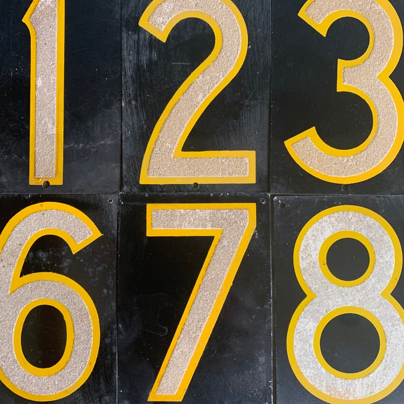 May include: Six black metal number tiles featuring the numbers 1, 2, 3, 6, 7, and 8. Each number has a textured, light-coloured centre with a yellow border. The tiles are arranged in two rows of three.
